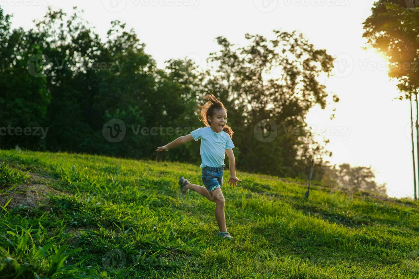 contento pequeño niña corriendo en el césped en el verano parque. pequeño niña corriendo mediante el prado con puesta de sol ligero. foto