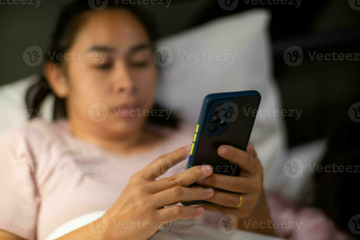 Young Asian woman lying on white bed and playing smartphone at night. Woman in bed with phone. Health and social concept. photo