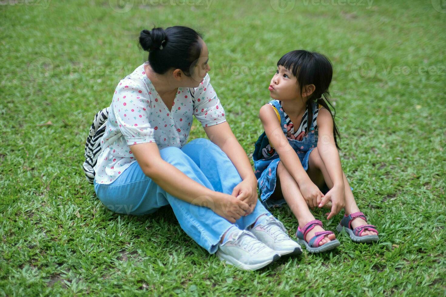 Mother and little daughter sitting on the grass together in the park. Mother having fun with her little daughter outdoors in green nature park. Happy family concept. Mother's Day photo