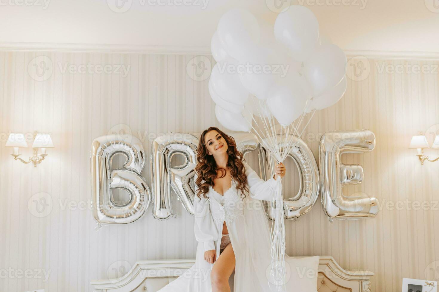 young curly girl with long hair in luxurious room standing on bed in white open dress holding many gel inflatable balls in hands and preparing for wedding ceremony photo