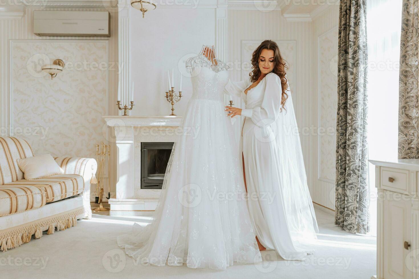 young curly girl with long hair in luxurious room standing near her dress on maeken white open dress getting ready for wedding ceremony photo