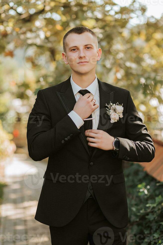 young groom fastens his tie looking into the lens outdoors. An adult man in a black suit and white shirt with a tie photo