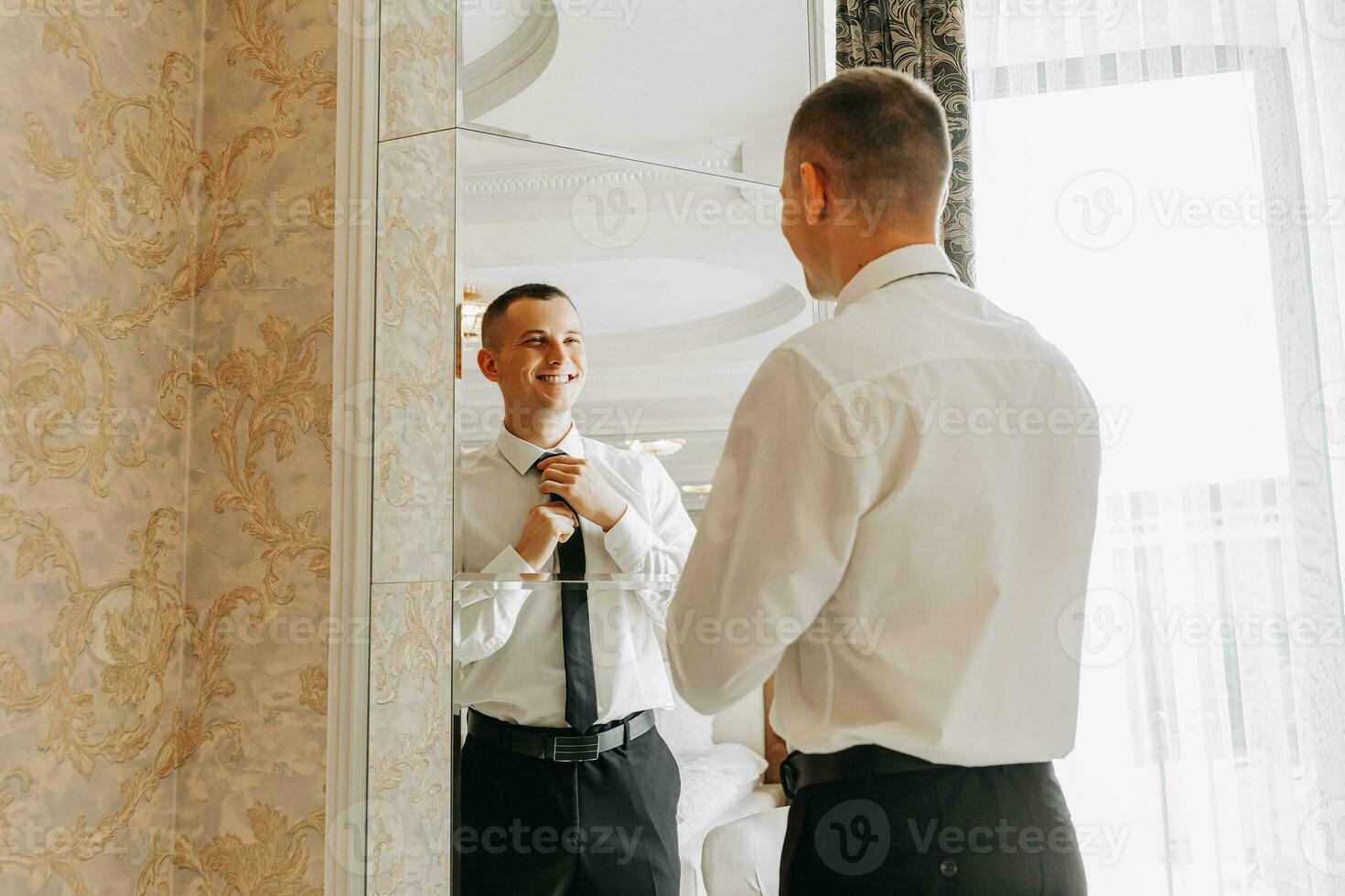 young man holding a tie in his hands in his room, near the hotel room mirror. The groom is preparing for the wedding ceremony photo