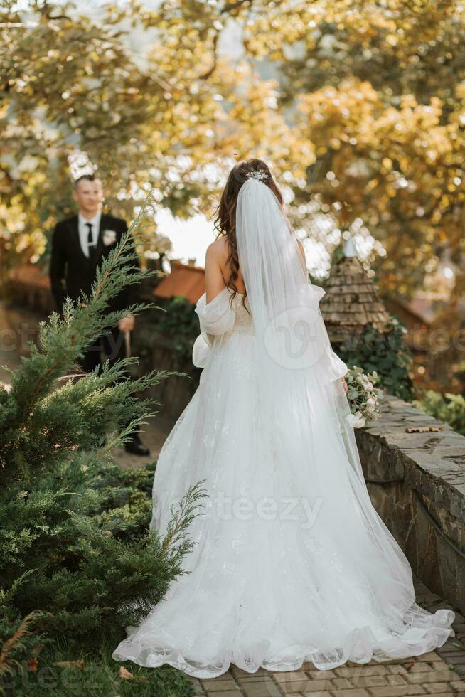 young and beautiful bride with long brown hair in a wedding dress outdoors with a wedding bouquet of flowers walking to her groom in the garden. Portrait from the back photo