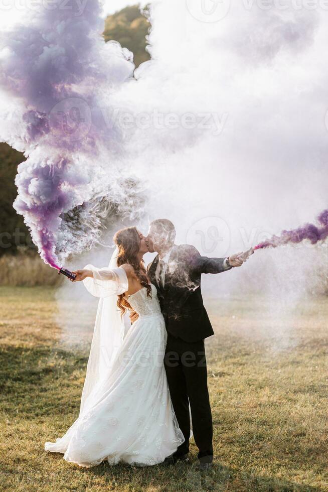 bride and groom playing with colored smoke in purple hands, hugs and kisses. Smoke bombs at a wedding. photo