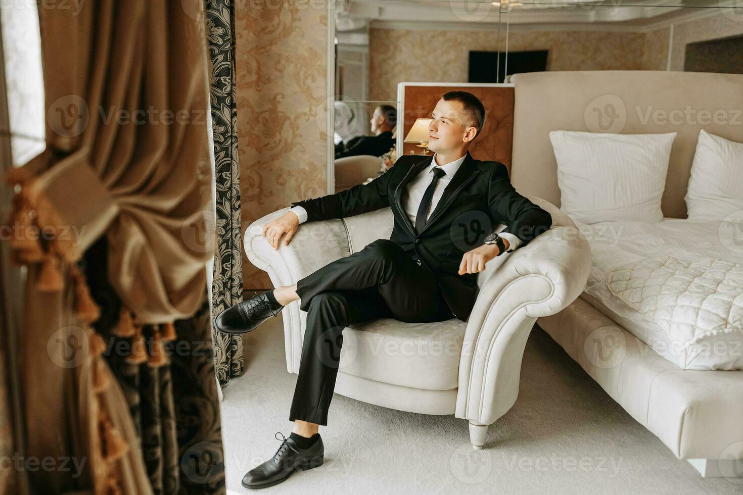 a young man is sitting in a chair in his room, near a window in a hotel room, a young businessman is wearing a black suit and a white shirt with a tie. The groom is preparing for the wedding ceremony. photo