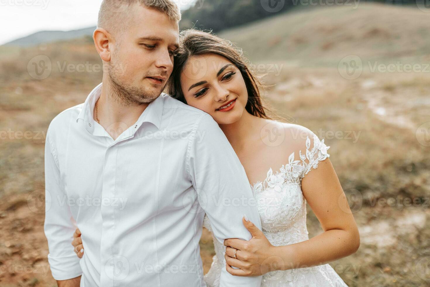 un elegante Pareja de europeo recién casados. un sonriente novia en un blanco vestir abrazos el novio desde detrás. el novio es vestido en un blanco camisa, sin un atar. al aire libre Boda en el montañas foto