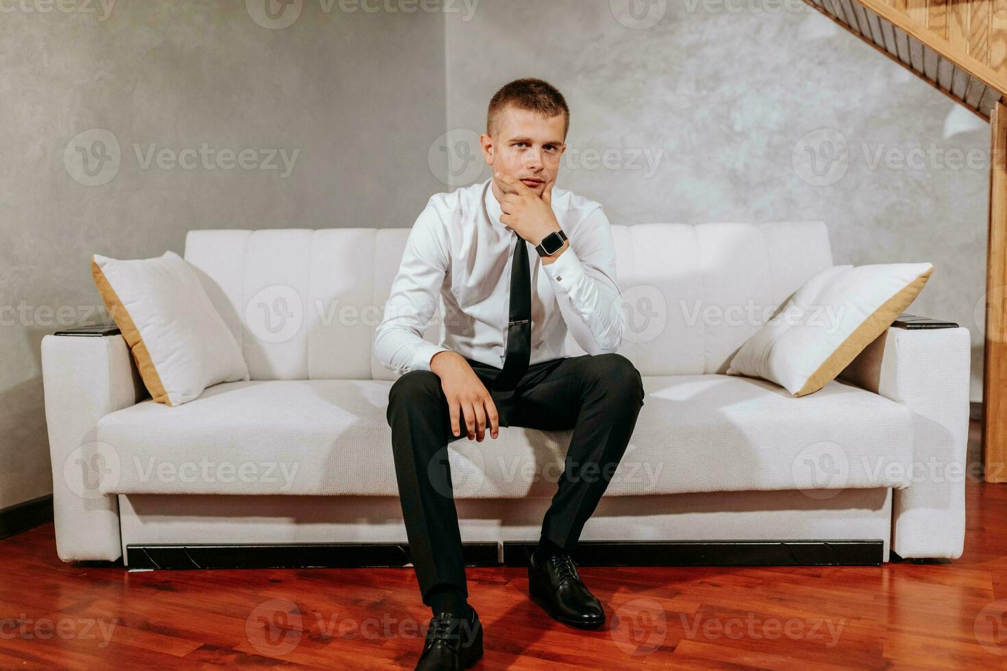 A mature man in a white shirt and black pants is sitting on a beige sofa in his room. Preparation for the wedding ceremony. Portrait of the groom. Free space photo