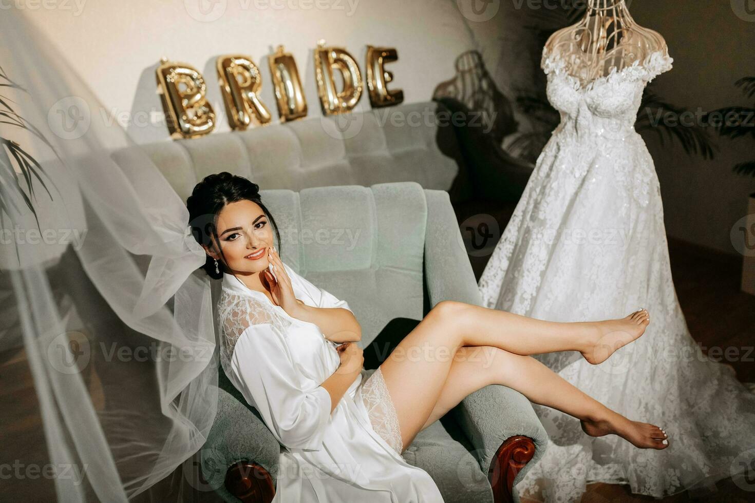 A grown-up bride sits on a chair in her room next to her wedding dress, with bare legs in a white robe. Gorgeous wedding hair and makeup photo