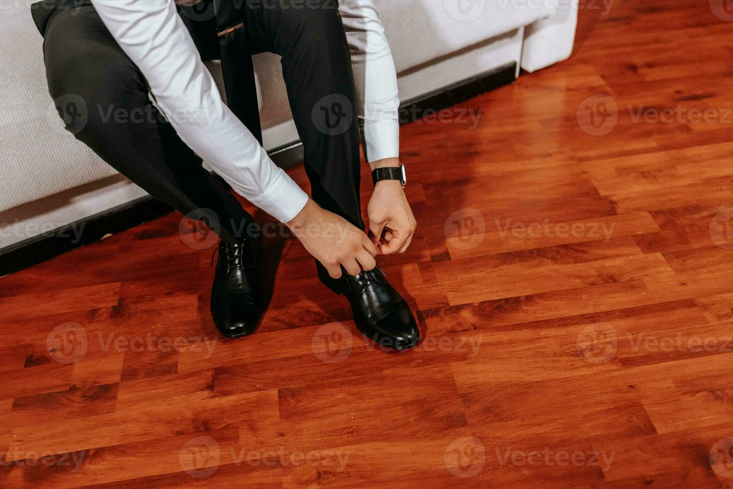 A mature man in a white shirt and black pants is putting on his shoes while sitting on the sofa in his room. Preparation for the wedding ceremony. Close-up photo. Free space photo