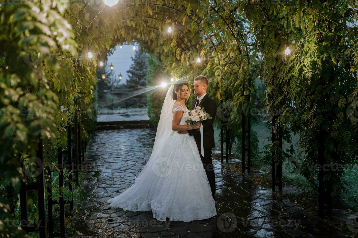 The bride and groom in the evening park with a bouquet of flowers in the middle of evening lights, greenery in nature. Romantic couple of newlyweds outdoors. Wedding ceremony in the botanical garden. photo