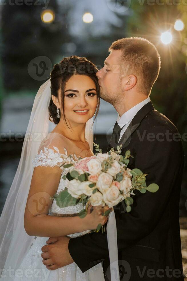 The bride and groom in the evening park with a bouquet of flowers in the middle of evening lights, greenery in nature. Romantic couple of newlyweds outdoors. Wedding ceremony in the botanical garden. photo