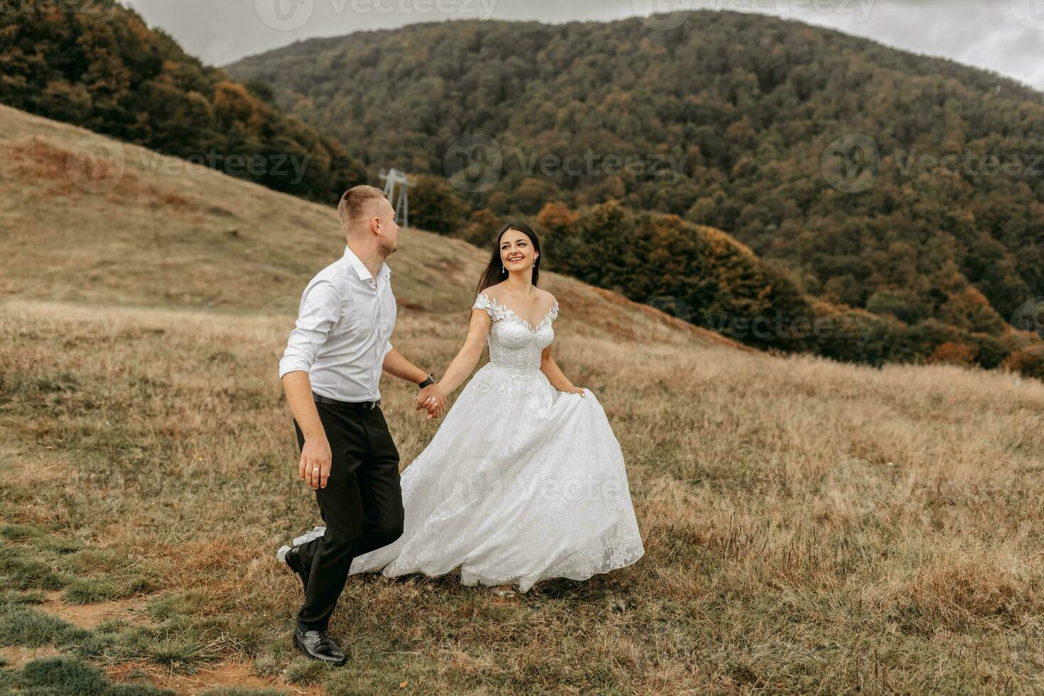 A romantic fairytale couple of newlyweds are running in a field at sunset, behind high mountains covered with trees. The bride in a white wedding dress, the groom in a white shirt and black pants photo