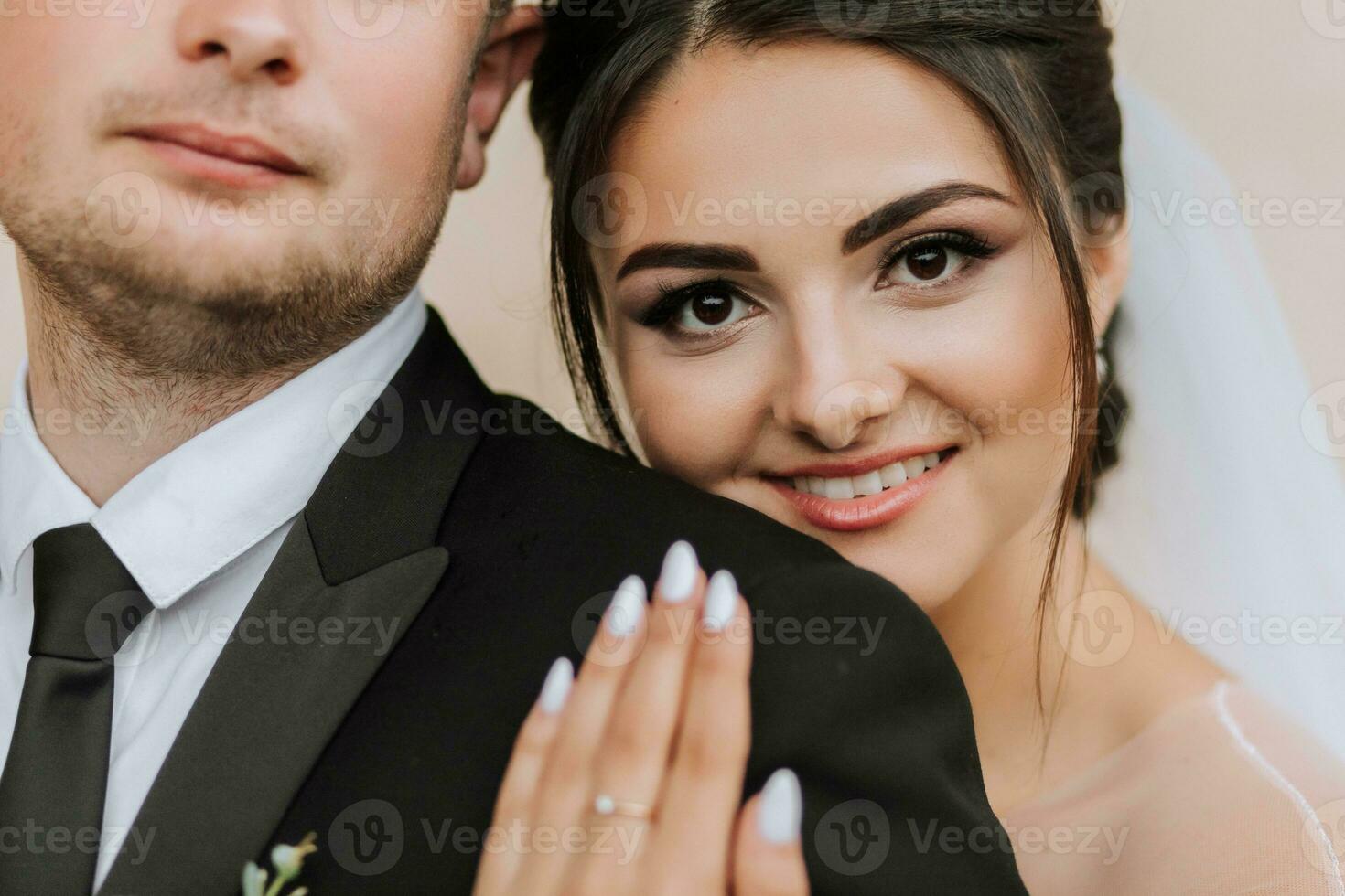 A stylish couple of European newlyweds. A smiling bride in a white dress hugs the groom from behind. Groom dressed in a classic black suit. Wedding in nature. Detailed portrait photo