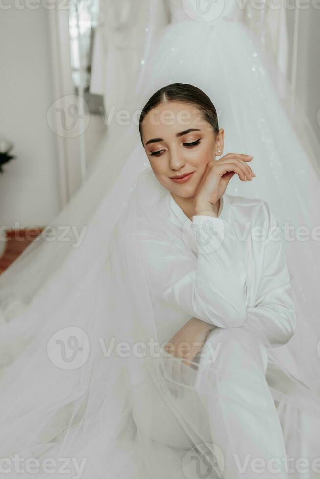 Portrait of a beautiful bride girl with stylish hair and makeup in a white robe, sitting on the train of her wedding dress, leaning on her hand photo