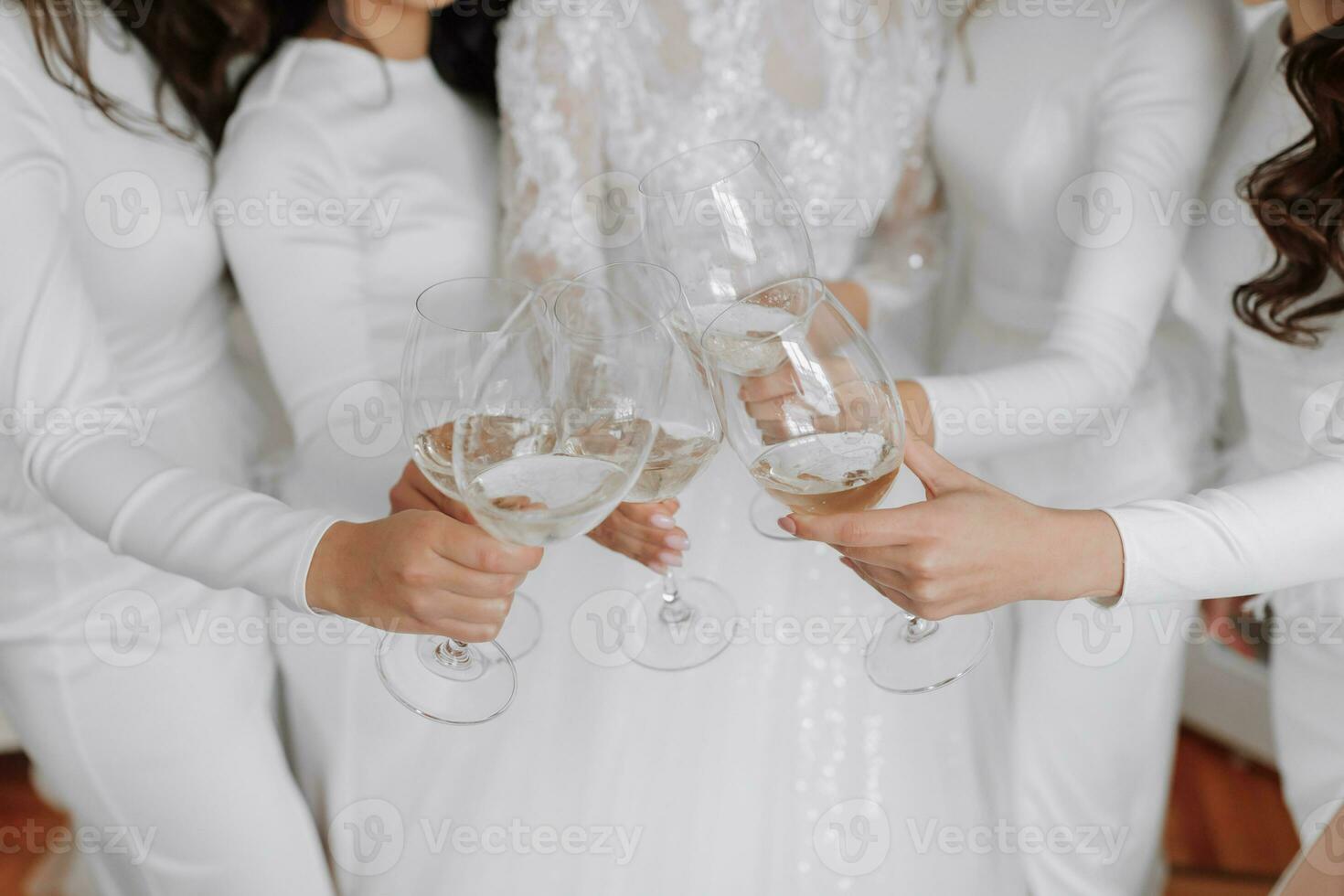 Young bridesmaids in white silk dresses drink champagne in the bride's room. Beautiful women celebrating bachelorette party standing in room and with champagne. close-up photo of hands with glasses