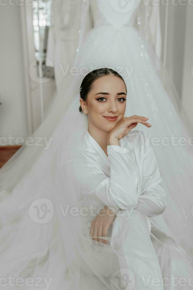 Portrait of a beautiful bride girl with stylish hair and makeup in a white robe, sitting on the train of her wedding dress, leaning on her hand photo