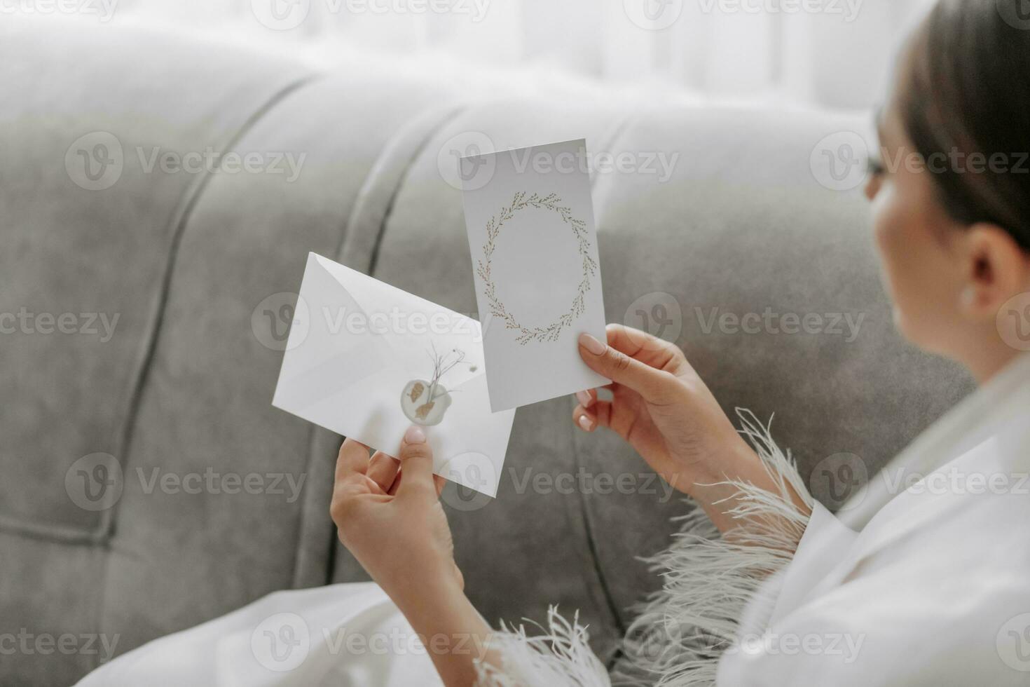 retrato de un hermosa niña novia con un elegante peinado y maquillaje en un blanco túnica, sentado en un sofá y participación un Boda invitación. gratis espacio foto