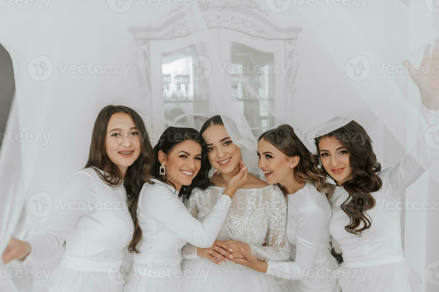 joven damas de honor en blanco seda vestidos abrazo y alegrarse en el de la novia habitación. hermosa mujer celebrando despedida de soltera fiesta en pie en habitación. foto