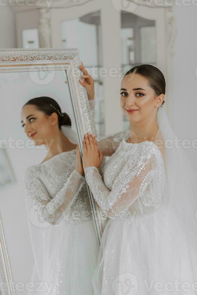 a beautiful bride in a wedding dress with closed shoulders stands by a mirror in her room, looks into the lens. reflection in the mirror photo
