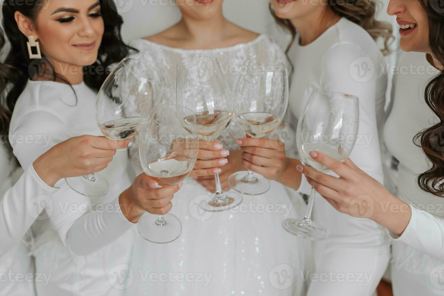 joven damas de honor en blanco seda vestidos bebida champán en el de la novia habitación. hermosa mujer celebrando despedida de soltera fiesta en pie en habitación y con champán. de cerca foto de manos con lentes