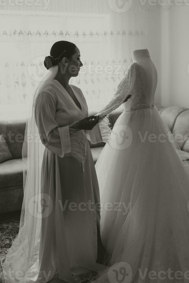 Stylish bride with wedding hairstyle and makeup in white robe standing near her wedding dress. Black and white photo