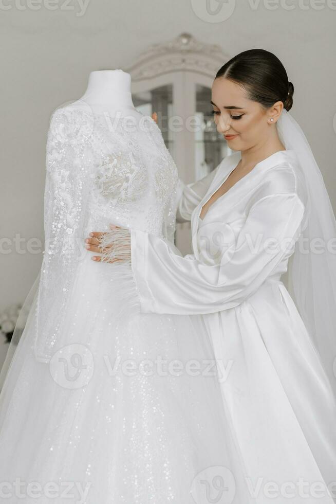 Stylish bride with wedding hairstyle and makeup in white robe standing near her wedding dress. photo