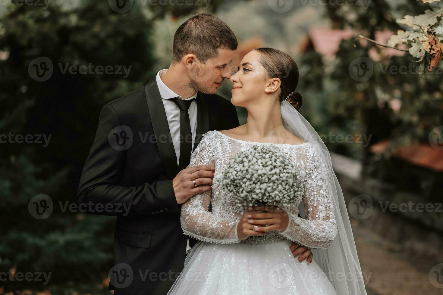 un elegante Pareja de europeo recién casados. un sonriente novia en un blanco vestir mira a el novio. el acicalar, vestido en un clásico negro traje, blanco camisa, . Boda en naturaleza foto