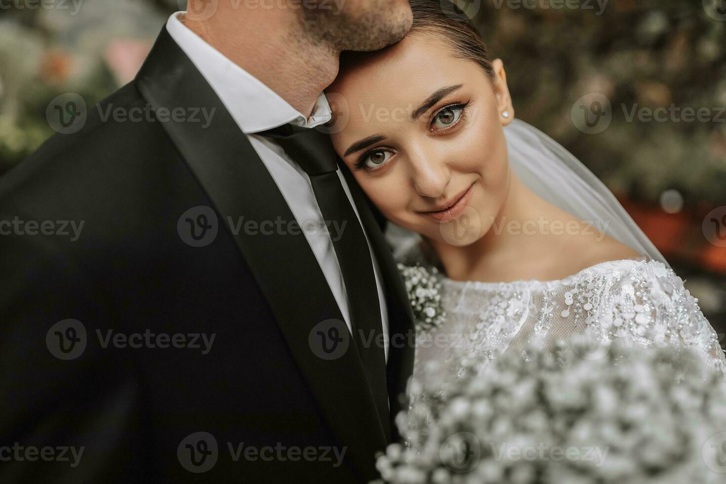 un elegante Pareja de europeo recién casados. un sonriente novia en un blanco vestir mentiras en el del novio hombro. el acicalar, vestido en un clásico negro traje, blanco camisa, . Boda en naturaleza foto