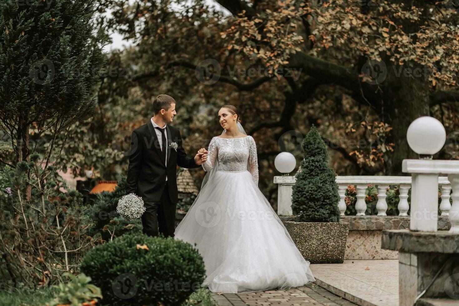 un elegante Pareja de europeo recién casados. sonriente novia en un blanco vestido. el novio es vestido en un clásico negro traje, blanco camisa y atar. Boda en naturaleza, un caminar en el jardín Entre alto arboles foto