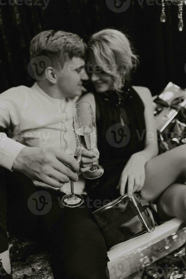 A romantic woman is lying in the bathroom with New Year's gifts, her husband is standing next to her with glasses of champagne. Merry Christmas. Black and white photo with artistic noises