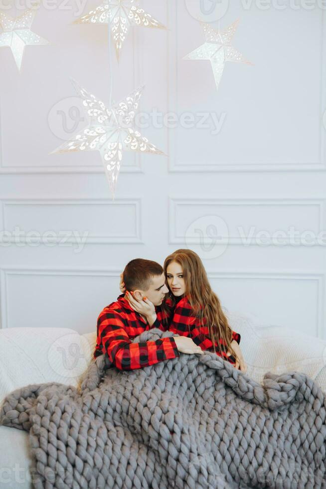 Young beautiful married couple in matching red pajamas sitting on a gray sofa, wrapped in a blanket, hugging and kissing. New Year's holidays and gifts under the Christmas tree photo