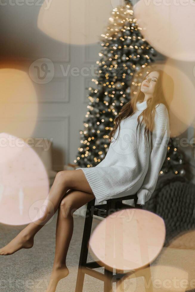 un hermosa y elegante joven niña en un blanco suéter poses en el Navidad luces en el antecedentes de el Navidad árbol foto