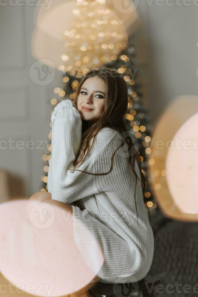A beautiful and stylish young girl in a white sweater poses in the Christmas lights on the background of the Christmas tree photo