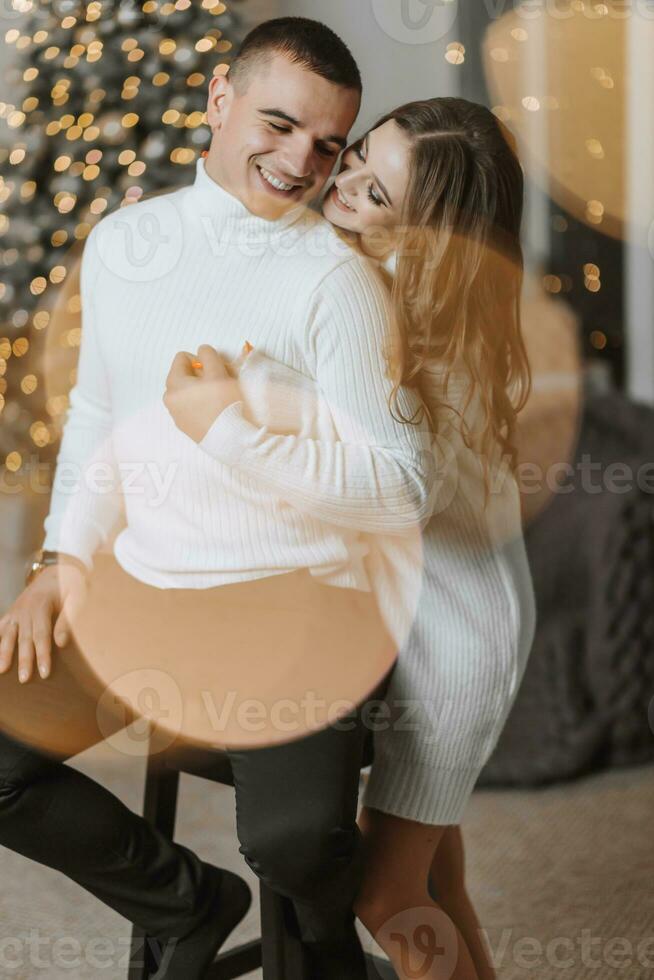 A beautiful and stylish young family in white sweaters hug and rejoice in the Christmas lights against the background of the Christmas tree. New year concept photo