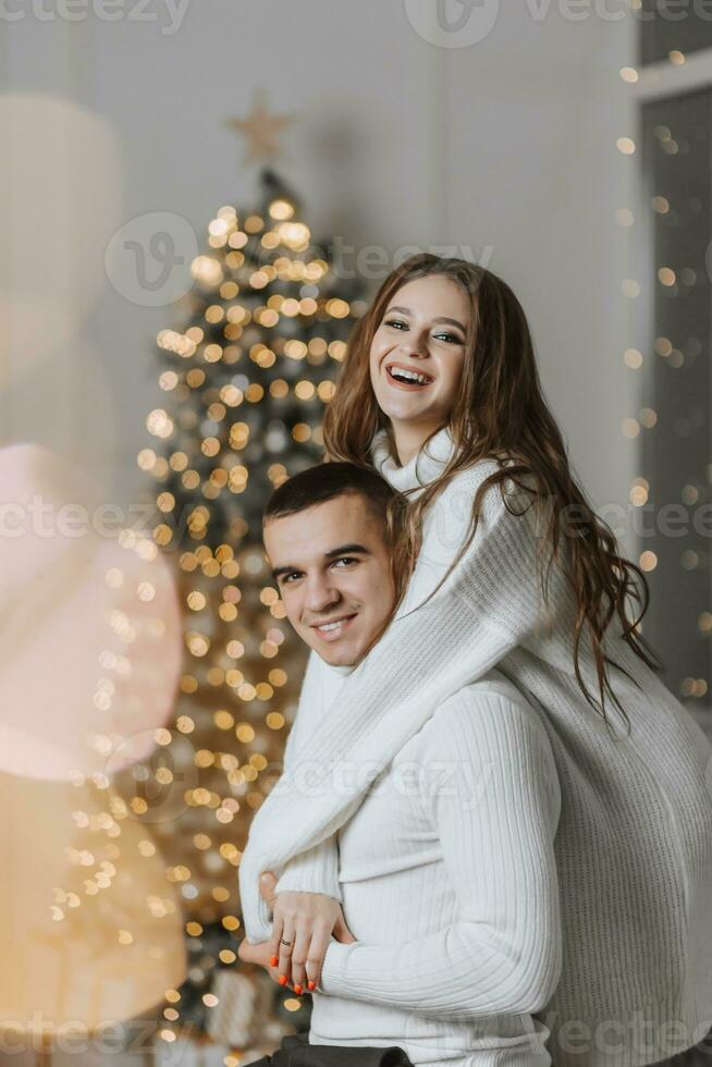 A beautiful and stylish young family in white sweaters hug and rejoice in the Christmas lights against the background of the Christmas tree. New year concept photo