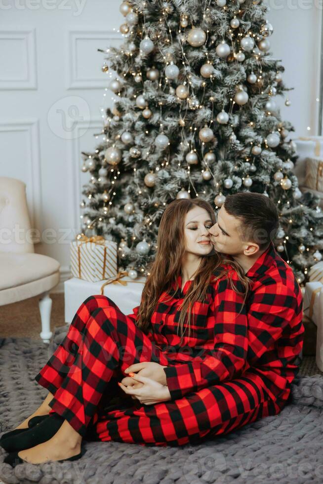 un hermosa joven niña en rojo pijama soportes en el antepecho y pone en un Navidad árbol. Navidad concepto. muchos regalos en papel envoltorios debajo el Navidad árbol foto