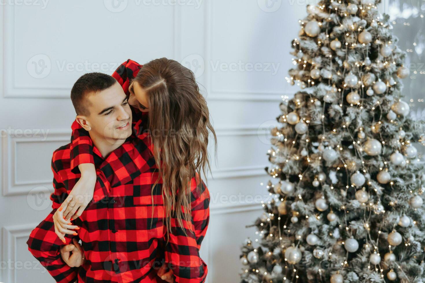 un hermosa joven niña en rojo pijama soportes en el antepecho y pone en un Navidad árbol. Navidad concepto. muchos regalos en papel envoltorios debajo el Navidad árbol foto