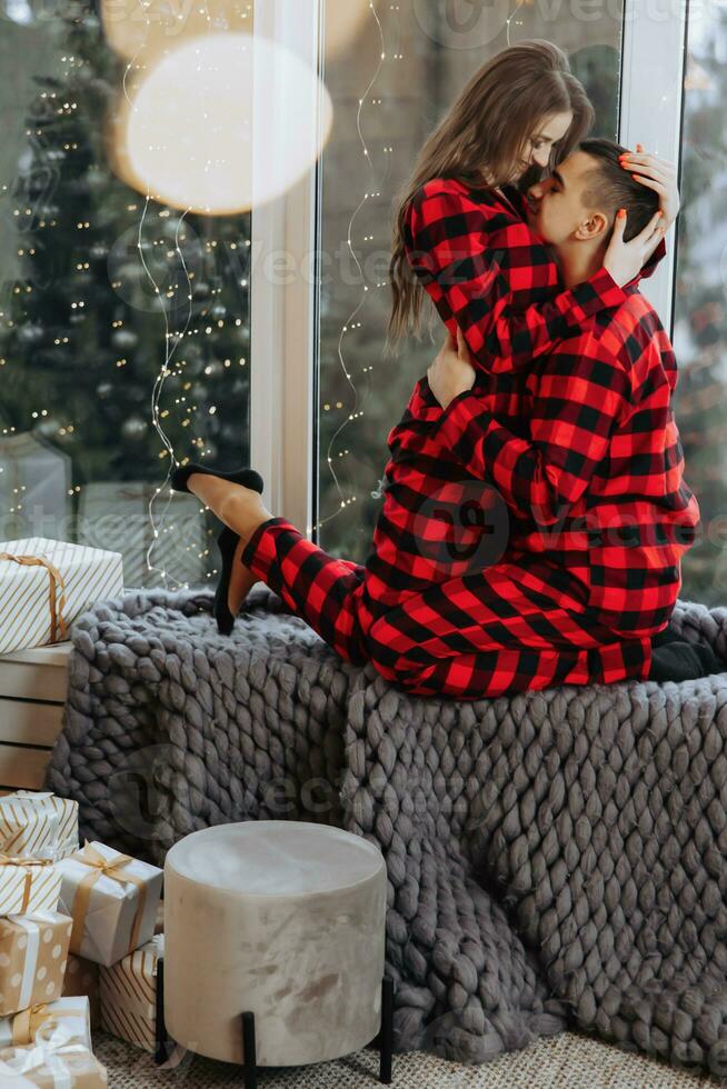 Young beautiful married couple in identical red pajamas on the windowsill hugging in the Christmas lights. Joy, hugs and kisses. New Year's holidays and gifts under the Christmas tree photo