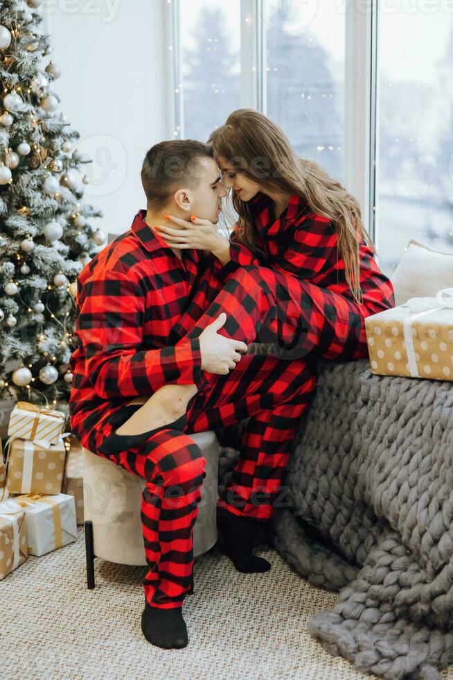 A young beautiful married couple in the same red pajamas, near the Christmas tree, are smiling, hugging and kissing. New Year holidays and gifts under the Christmas tree photo