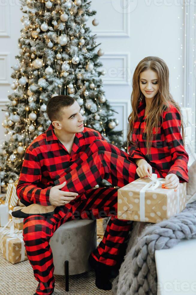 A young beautiful family couple in the same red pajamas, near the Christmas tree, rejoice and exchange New Year's gifts. New Year holidays and gifts under the Christmas tree photo