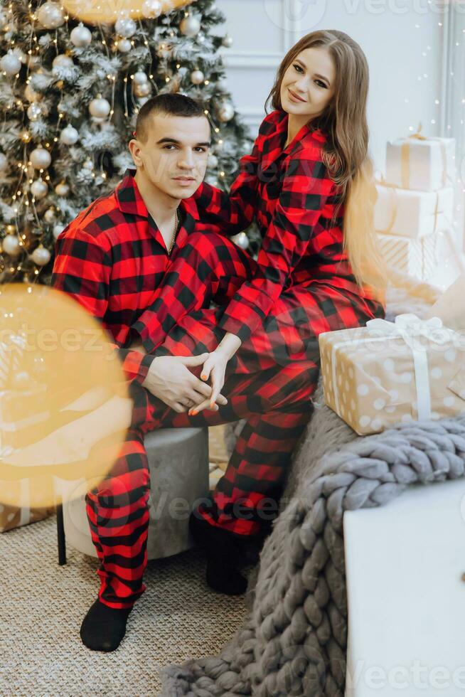 A young beautiful family couple in the same red pajamas, near the Christmas tree, rejoice and exchange New Year's gifts. New Year holidays and gifts under the Christmas tree photo