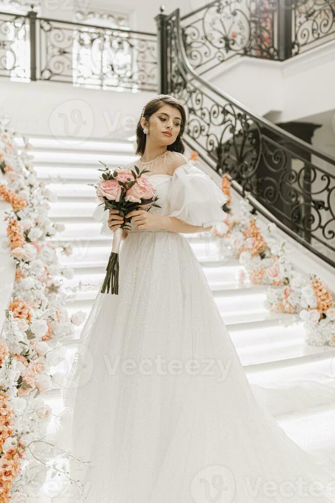 full-length portrait of a young girl with beautiful hair and makeup in a luxurious wedding dress on the steps of a restaurant near rose flowers. Vertical photo. photo