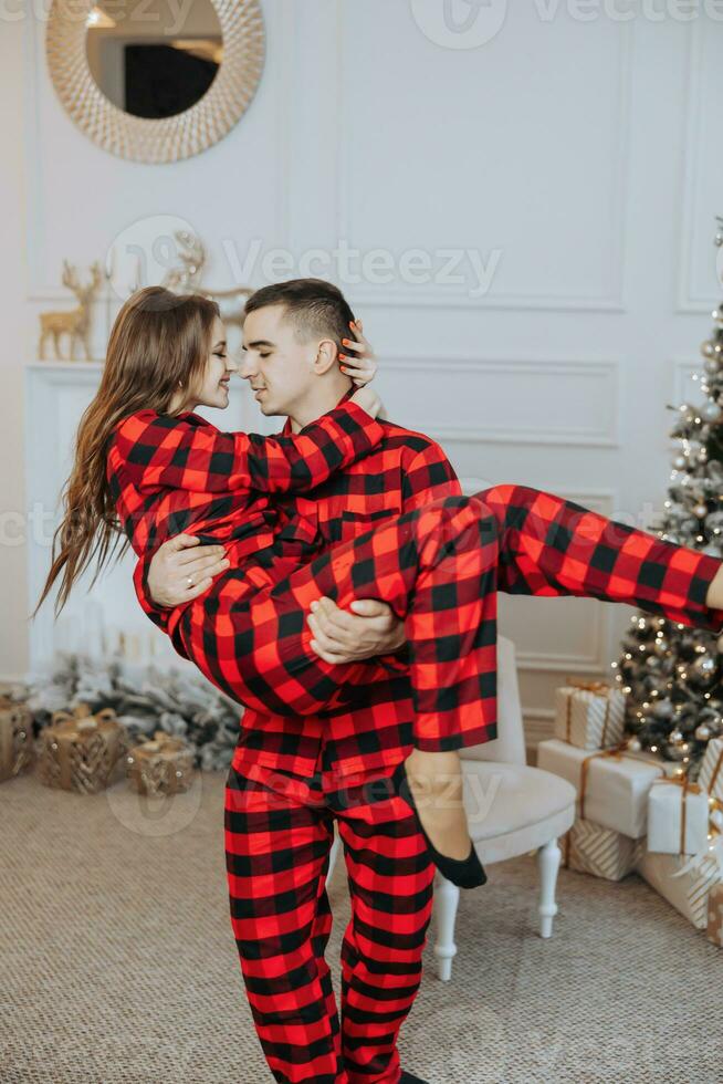 a beautiful young girl in red pajamas stands on the windowsill and puts on a Christmas tree. Christmas concept. Many gifts in paper wrappers under the Christmas tree photo