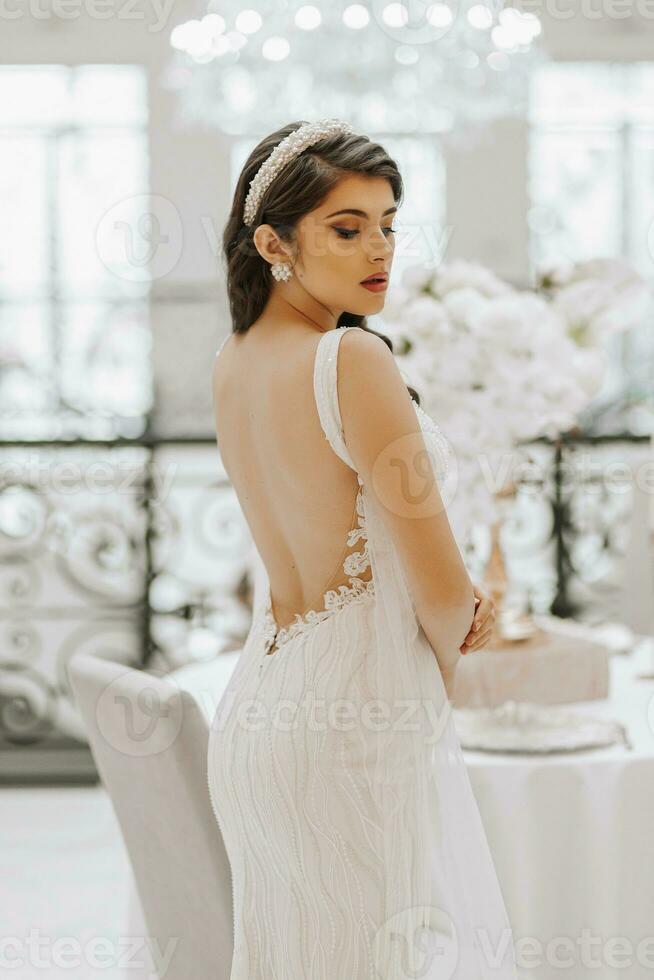 Beautiful bride at the table with a vase of flowers. The woman is wearing a long wedding dress with lace and an open back, a bow is tied on the back. photo