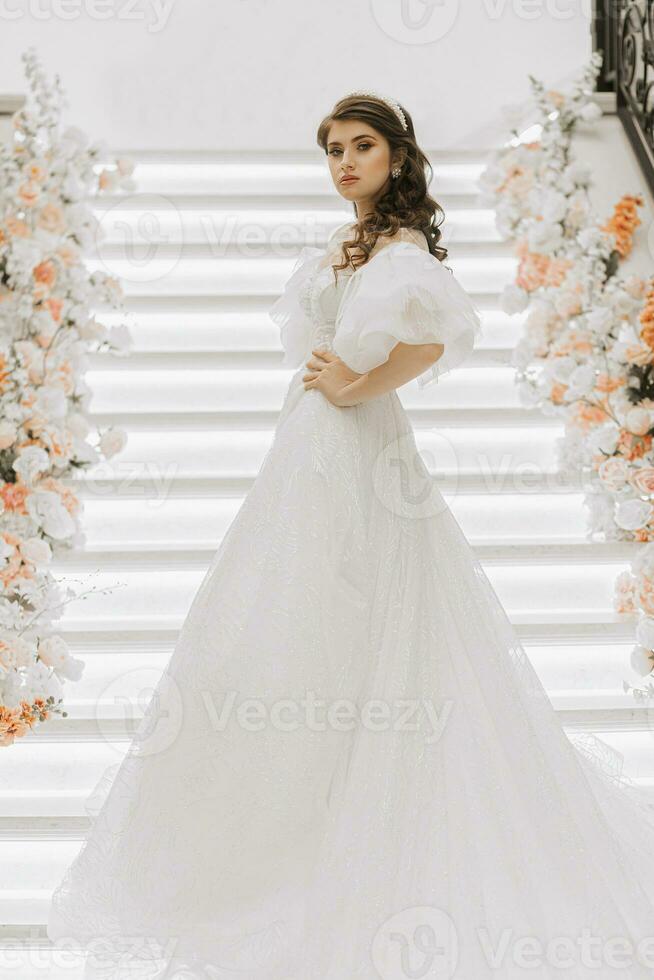 full-length portrait of a young girl with beautiful hair and makeup in a luxurious wedding dress on the steps of a restaurant near rose flowers. Vertical photo. photo