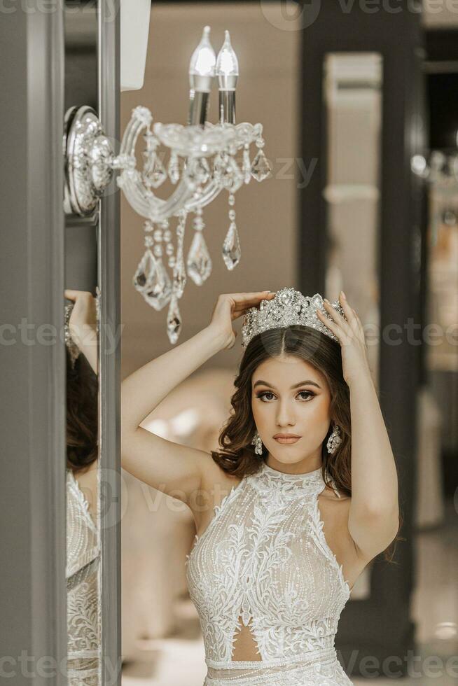 Portrait of a girl in a wedding dress near a mirror, with a crown on her head. The concept of a royal feast. Vertical photo. photo