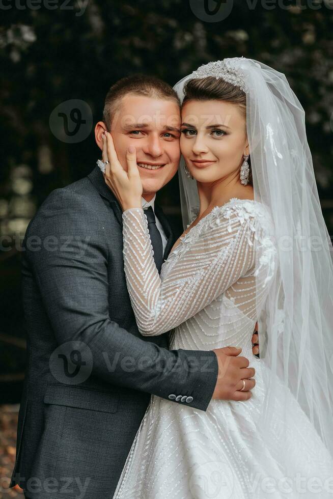 Boda retrato. un morena novia en un largo vestir y un novio en un clásico traje son de pie, abrazando, en amor y posando para el cámara. hermosa novia, cordón vestir foto