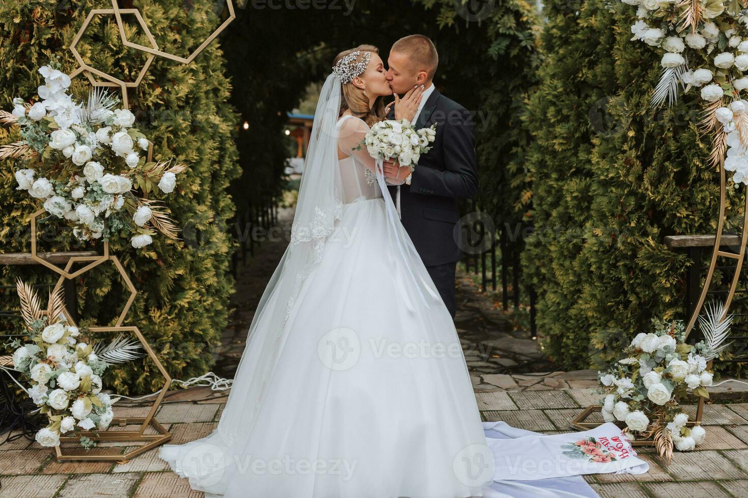 un joven Boda Pareja a un Boda pintura ceremonia. el novia y novio Beso en contra el antecedentes de verde arboles y un arco decorado con flores foto