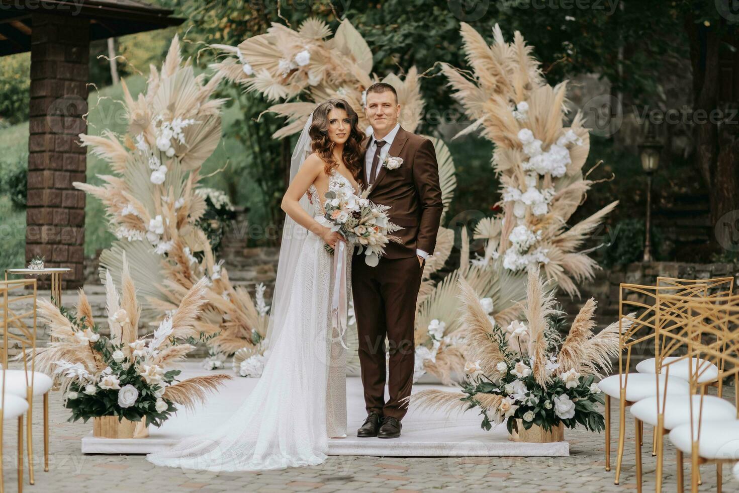 novio y novia en el antecedentes de el Boda arco. recién casados con un Boda ramo de flores en pie a un Boda ceremonia debajo un arco decorado con flores y seco flores al aire libre. foto